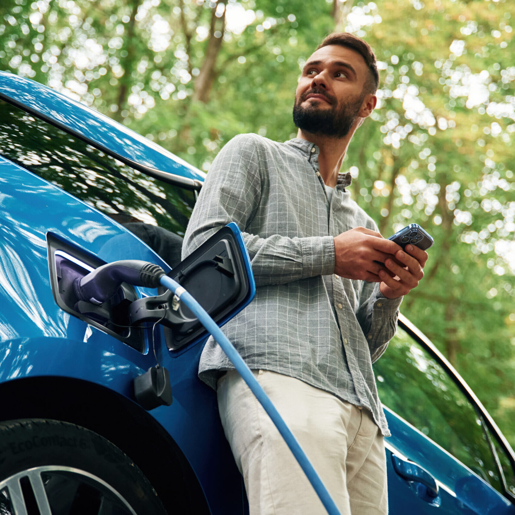 Trees behind. A handsome man is standing outdoors near an electric automobile and waiting for charging.